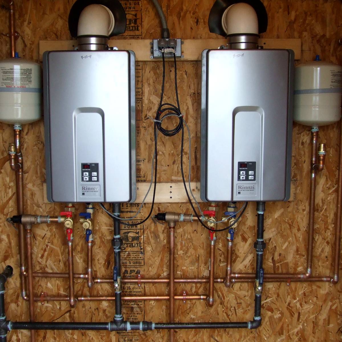 Two gas tankless water heaters mounted side by side in a garage, with expansion tanks, isolation valves and copper supply lines neatly run along the wall